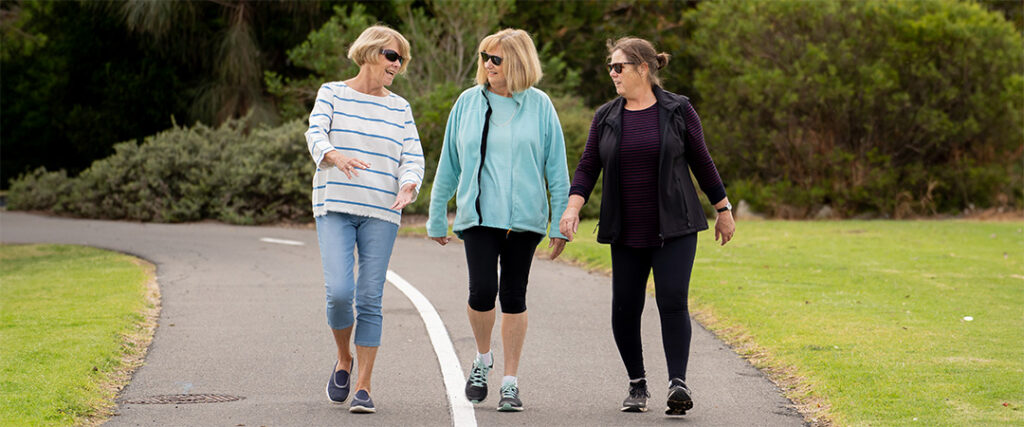 Three women wearing casual clothes and sunglasses walk together on a paved path in a park, talking and smiling. Green grass, bushes, and trees line the path.